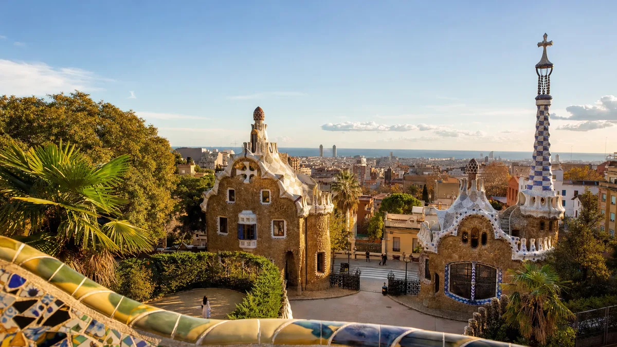 AI-generated image: Park Güell, Barcelona: A sunlit view of the vibrant mosaics and whimsical structures of Park Güell, surrounded by lush greenery. In the background, a few visitors stroll leisurely